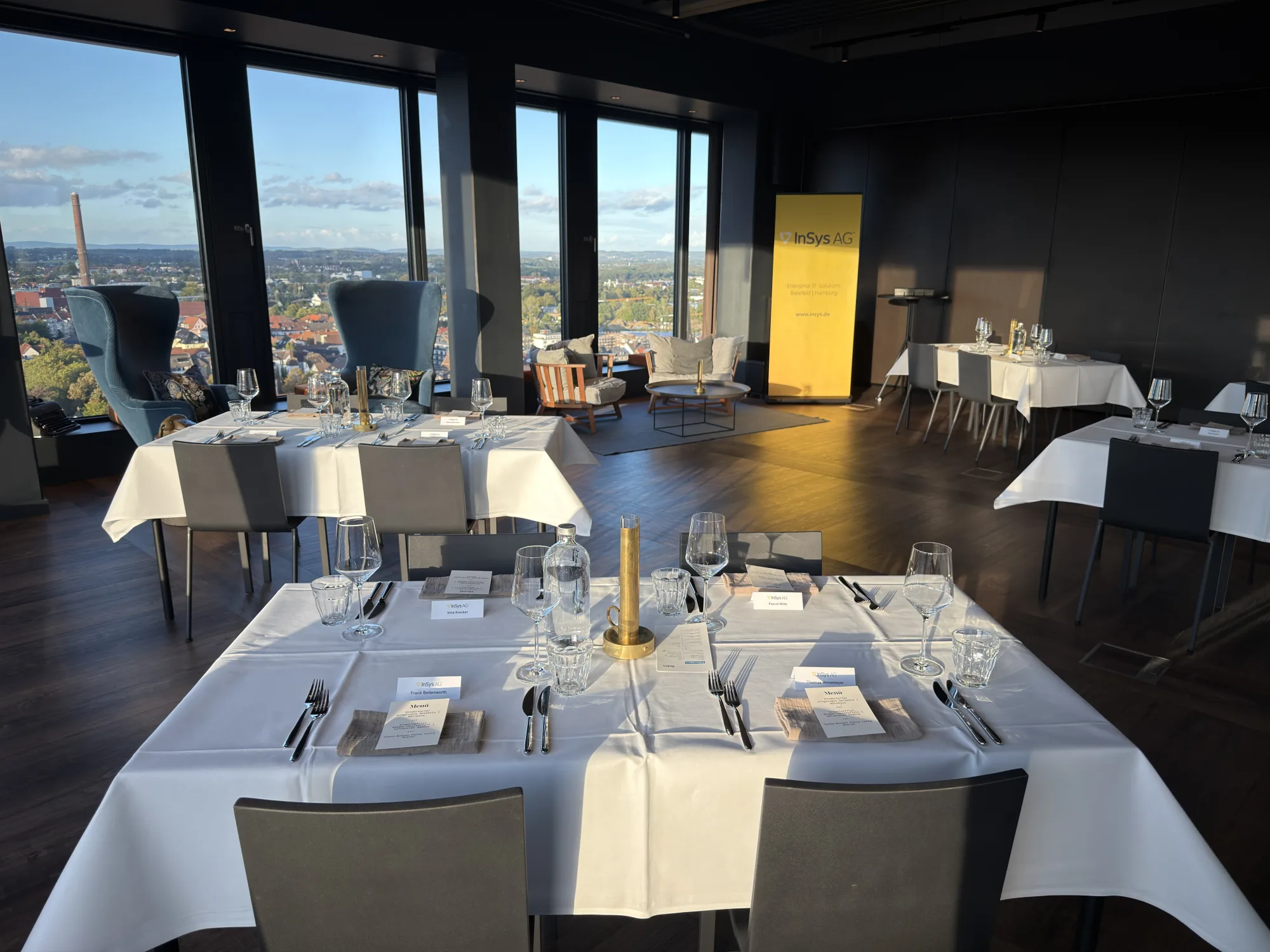 Gedeckte Tische mit weißen Tischdecken und Platzkarten in der Skybar des H1 Bielefeld zur InSys AG® Connect 2026, dahinter ein Roll-up und der Blick über Bielefeld